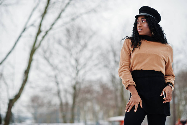 African american woman in black skirt, brown sweater and cap posed at winter day against snowy background, holding phone at hand. - Foto, Bild