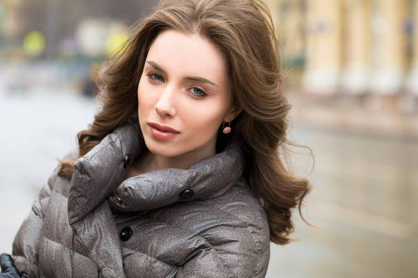 Closeup portrait of a young beautiful brunette woman in the winter down jacket on the background of cloudy street - Photo, image