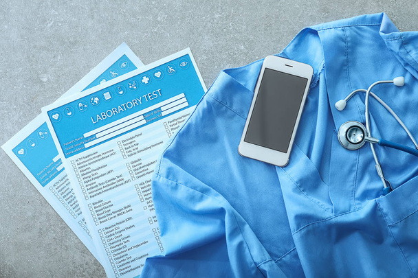 Stethoscope with mobile phone, doctor's uniform and lists of laboratory tests on grey table. Health concept - Photo, Image