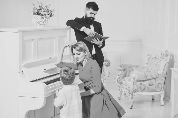 Child stand near piano keyboard, white interior background. Musician education concept. Mother teaches kid play piano, musical instrument. Boy adorable try to play piano, while parents watching him - Φωτογραφία, εικόνα