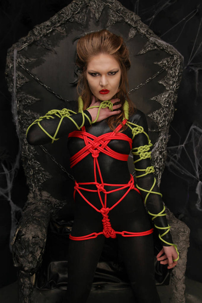 beautiful young girl standing in a dark fetish spandex catsuit tied with colorful ropes in shibari art style - Photo, Image