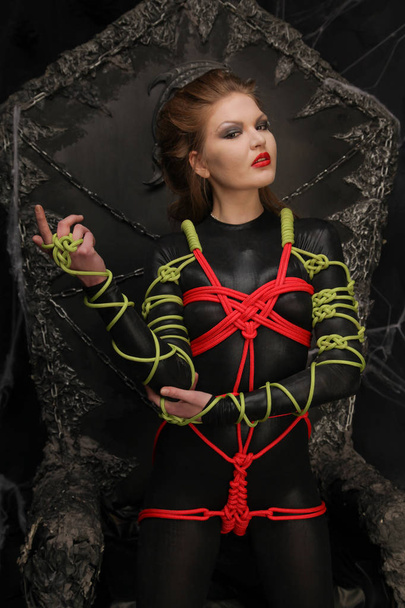 beautiful young girl standing in a dark fetish spandex catsuit tied with colorful ropes in shibari art style - Photo, Image