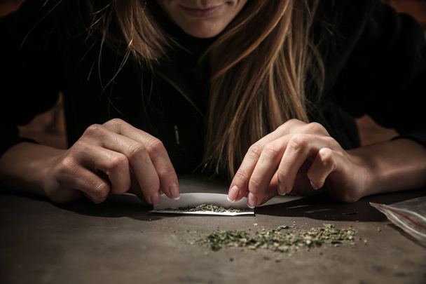 Female junkie making cigarette with marijuana at grey table. Concept of addiction - Φωτογραφία, εικόνα