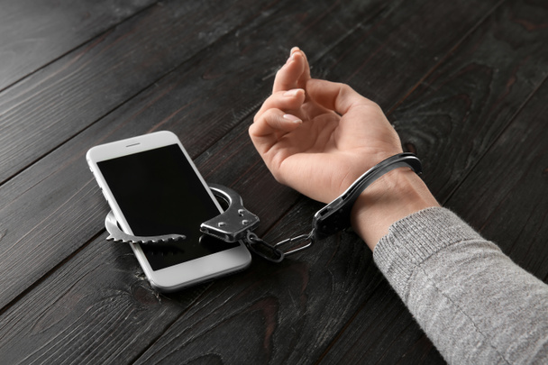 Female hand in handcuffs with mobile phone on wooden background. Concept of addiction - Fotoğraf, Görsel