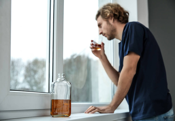 Depressed young man drinking whiskey near window. Alcoholism concept - Photo, Image