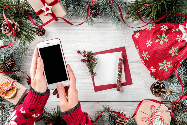 Cep telefonu tutan eller. Noel hediye kutuları kırmızı dekorasyon, çam dalları, çam kozalakları, kağıt kartı kayda göre tatil arka plan ile. Noel ve mutlu yeni yıl kartı. Düz yatıyordu, en iyi görünümü - Fotoğraf, Görsel
