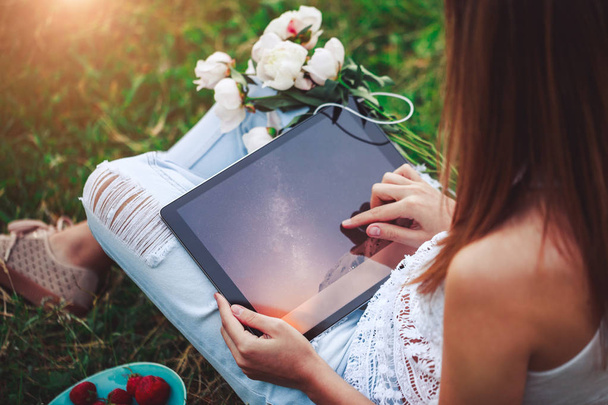 Молода жінка використовує планшетний ПК, сидячи на літній траві з букетом квітів півоній. Літні канікули
 - Фото, зображення