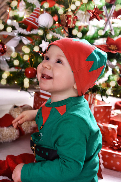 little boy (10 mo) sitting in the room in front of christmas tree and enjoing his first christmas  - Foto, afbeelding