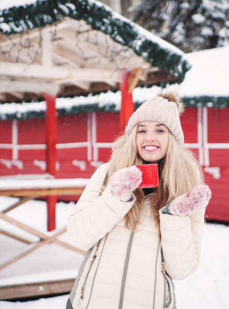 Smiling beautiful young woman drinks a hot cocoa outdoor. Snowy weather. Winter holidays. - Zdjęcie, obraz