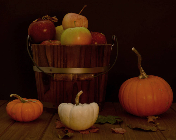 Balde de maçãs sortidas contra fundo preto na mesa de madeira decorada com abóboras laranja e branca e algumas folhas de queda composição queda
  - Foto, Imagem