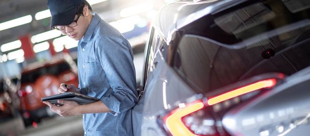 Молодой азиатский автомеханик держит цифровой планшет, проверяя автомобиль в гараже автосервиса. Инженер по техническому обслуживанию, работающий в автомобильной промышленности. Концепция технического обслуживания и ремонта автомобилей
 - Фото, изображение