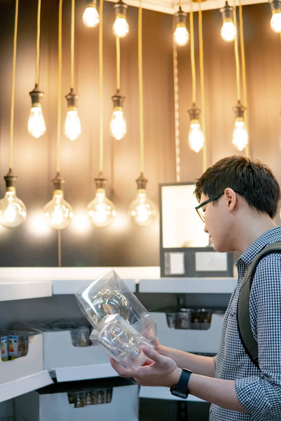 Jeune homme asiatique choisissant paquet d'ampoule électrique dans le magasin. Meubles décoration à domicile concept shopping
 - Photo, image