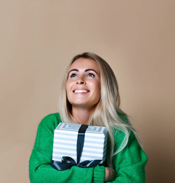 Portrait of a pretty smiling blonde girl holding gift box and looking at it, isolated over color beige background - Foto, Bild