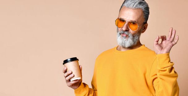 fashionable man senior looking at camera, having cap with coffee in arms and shows a sign ok with his fingers - Foto, Imagen