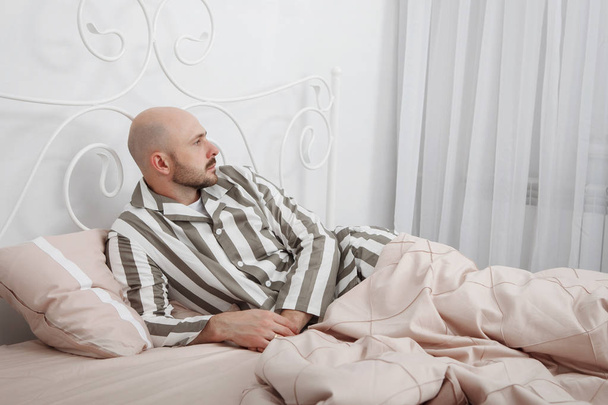 Guapo calvo, barbudo hombre en pijama a rayas está acostado y pensando en la cama con almohadas y ropa de cama rosa
. - Foto, imagen