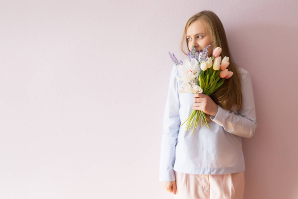 A young beautiful girl with blond dissolved long hair, a felt hat on her head, keeps spring flowers in her hands on a sunny day. Women's Day. - Foto, imagen