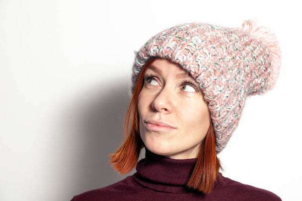 Isolated portrait of beautiful young redhead girl with green eyes looking sideaways in purple sweater and pink knitted hat with pompon dressed sideways smiling on white background in studio - Zdjęcie, obraz