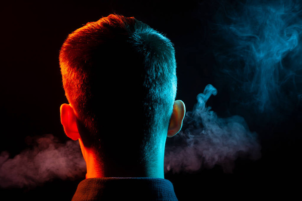 Вид ззаду на голову хлопчика в сорочці, що курить вейп і видихає різнокольоровий дим зеленого і червоного кольорів в різних напрямках від себе на чорному ізольованому фоні
 - Фото, зображення