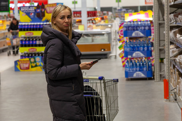 ein junges blondes Mädchen in langer Jacke wählt beim Einkaufen im Einkaufszentrum mit Einkaufswagen und Telefon in der Hand in der Bäckereiabteilung zwischen einer großen Anzahl von Regalen und Theken - Foto, Bild