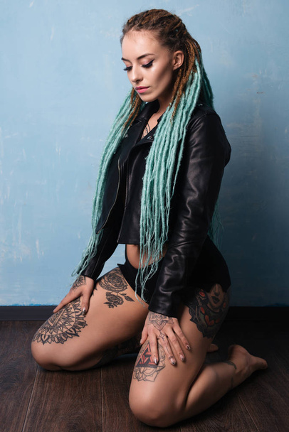 Portrait of a girl with tattoos and dreadlocks dressed in a leather jacket sits on her lap on the floor - Foto, Imagem