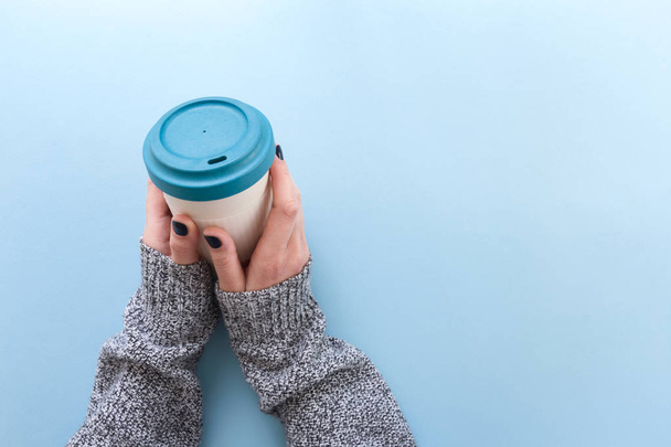 Hands in long sleeve knitted sweater, holding bamboo reusable cup with lid on, overhead on blue background. - Fotó, kép