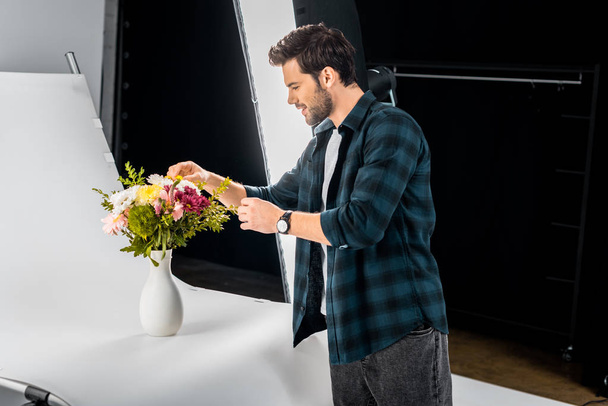 joven fotógrafo sonriente arreglando flores en estudio fotográfico
  - Foto, Imagen