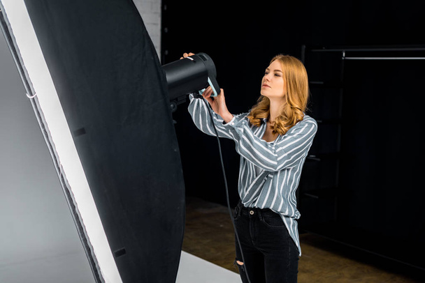 молодая женщина-фотограф, работающая с осветительным оборудованием в фотостудии
 - Фото, изображение