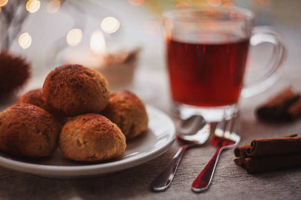 Three little curd and oatmeal buns with a cup of black tea, decorated with cinnamon, Christmas lights and hand made candle in background - Photo, Image