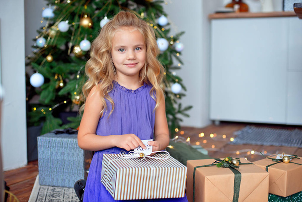 Little beautiful girl with blond curly hair considers her gifts against the background of the Christmas tree. Christmas gift, New Year's gift Christmas concept. - Photo, Image