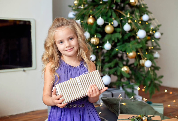 A beautiful girl with blond curly hair stands in the living room near the fireplace and holds her gift in her hands. New Year's concept. - Foto, Bild