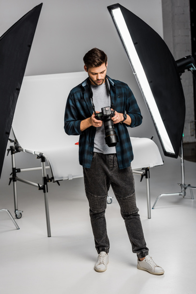 full length view of handsome professional young photographer using camera in photo studio  - Photo, Image
