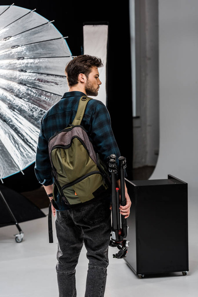 back view of young man with backpack holding professional equipment in photo studio  - Фото, изображение