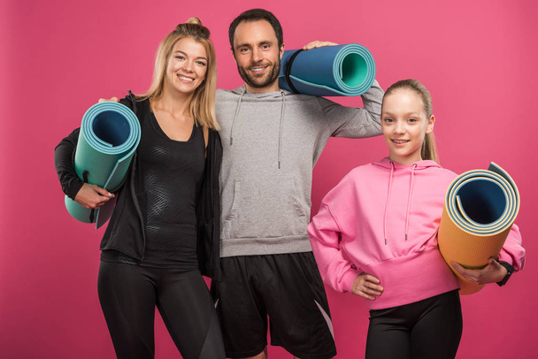 athletic mother, father and daughter holding fitness mats, isolated on pink - 写真・画像
