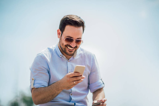 Sonriente joven hombre casual sosteniendo el teléfono móvil y la tarjeta de crédito mientras está sentado al aire libre
  - Foto, Imagen
