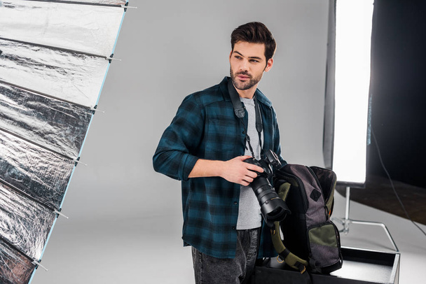 handsome professional young photographer standing with photo camera and looking away in studio - Foto, immagini