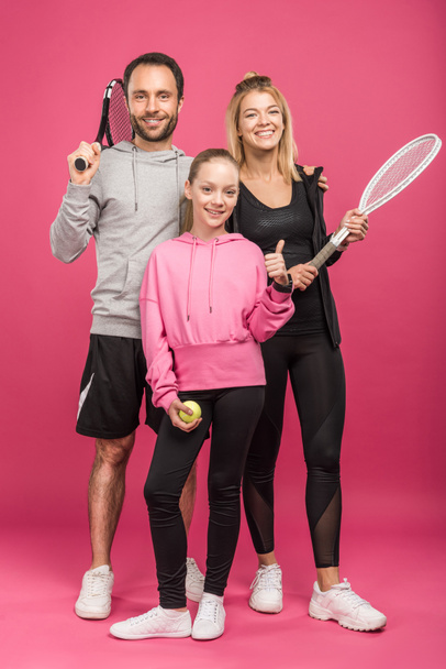sportive family holding tennis rackets and ball, while child showing thumb up, isolated on pink - 写真・画像