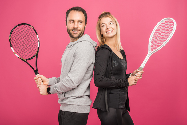 happy athletic couple holding tennis rackets, isolated on pink - Фото, изображение