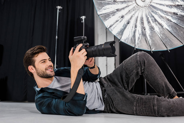 jeune photographe souriant couché et utilisant un appareil photo en studio
 - Photo, image