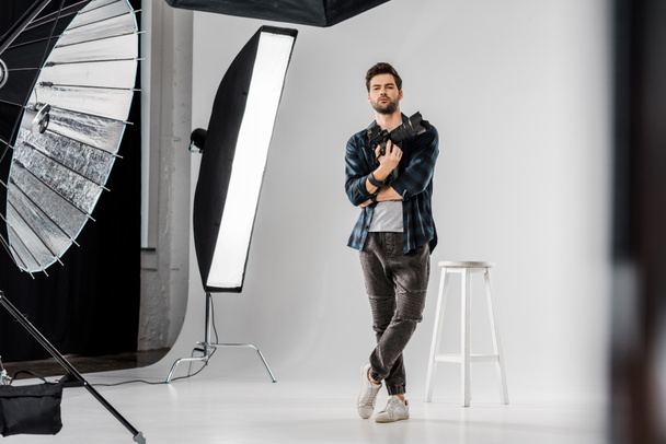 full length view of confident young photographer holding professional camera and looking at camera in photo studio  - Foto, immagini