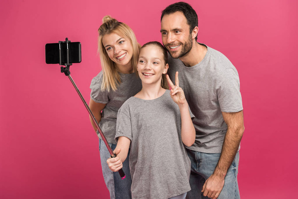 smiling family taking selfie on smartphone while daughter showing peace symbol, isolated on pink - 写真・画像
