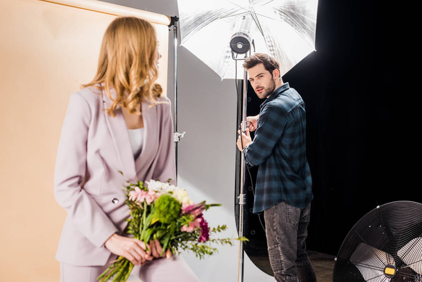 photographer working with lighting equipment and looking at young female model with flowers in studio  - Valokuva, kuva