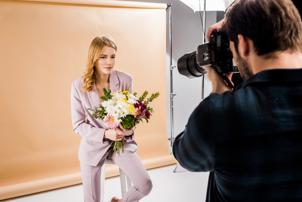 φωτογράφος γυρίσματα όμορφη νεαρή γυναίκα που ποζάρει με λουλούδια στο στούντιο - Φωτογραφία, εικόνα