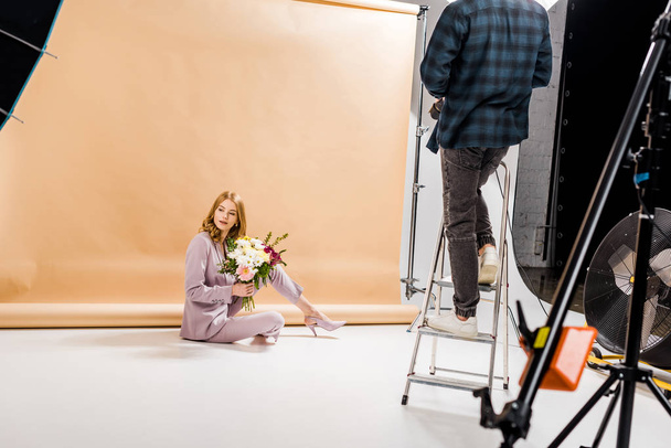 cropped shot of photographer standing on step ladder while beautiful girl posing with bouquet of flowers in studio   - Photo, Image
