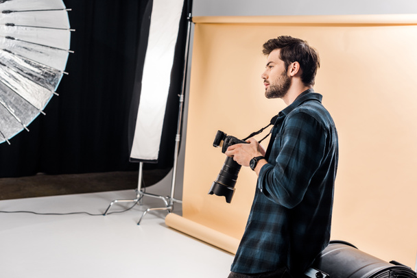 vue latérale du beau jeune photographe tenant la caméra et regardant loin dans le studio de photo
 - Photo, image