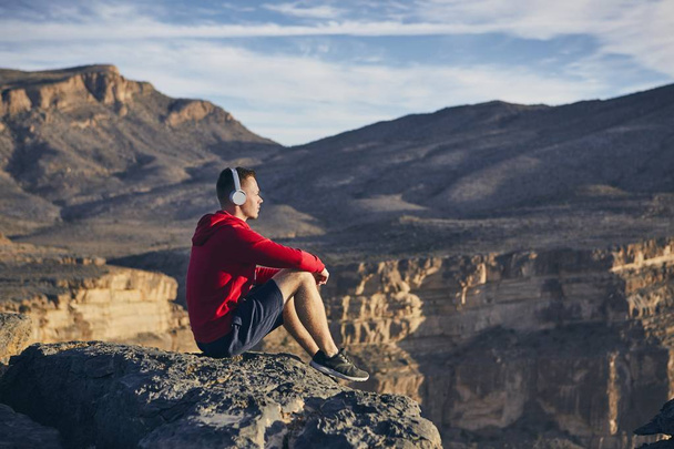Relaxation in mountains. Young man with headphones sitting on the edge of cliff and listening music. Jebel Akhdar, Grand Canyon of Oman.  - Fotoğraf, Görsel