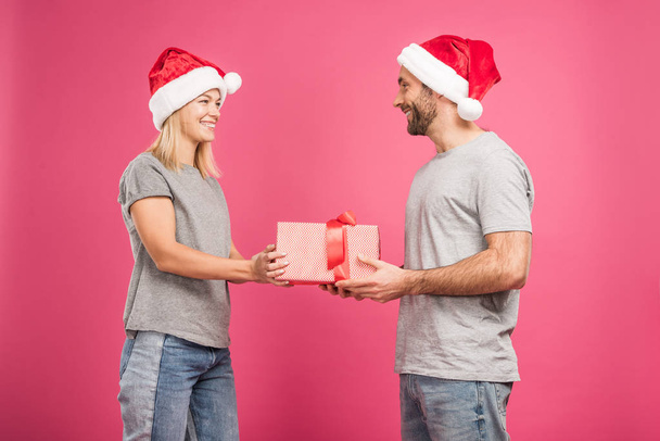 beautiful cheerful couple in santa hats gifting christmas present, isolated on pink - Photo, Image