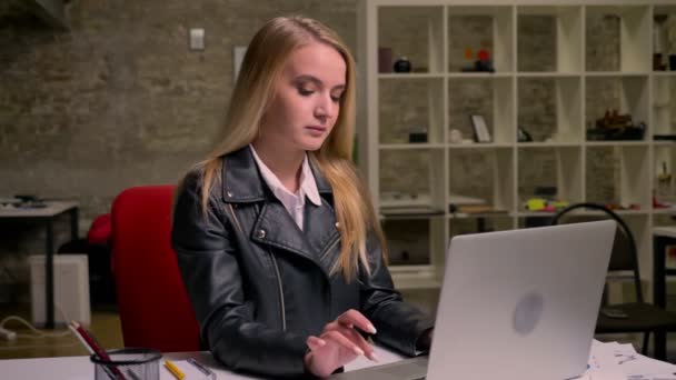 Beautiful blonde caucasian woman is typing on laptop focused and then looking at camera and smiling while being in light brick office, working vibes - Footage, Video