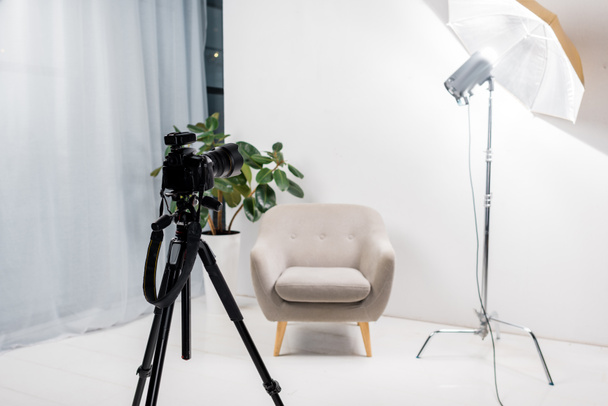 close-up view of professional photo camera on tripod, empty armchair and lighting equipment in studio - Foto, afbeelding