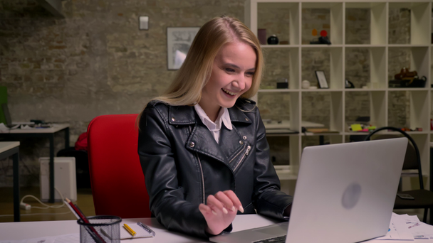 Enjoyable caucasian blonde female is laughing and relaxed looking at computer screen whily typing on keyboard, hanging in light office at workplace - Footage, Video