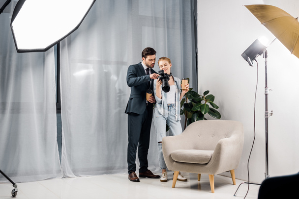 female photographer and young businessman holding paper cups and checking photos on camera in studio - Fotó, kép
